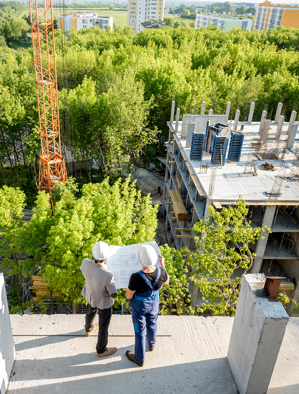 2 travailleurs du bâtiment, tenant un plan devant un immeuble en construction, vue du dessus, immeuble en construction en contre-bas