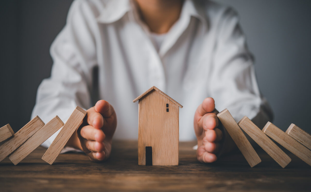 Maison miniature en bois protégée par des mains face à des dominos, symbolisant la sécurité de l’investissement immobilier locatif en LMNP et la gestion des risques